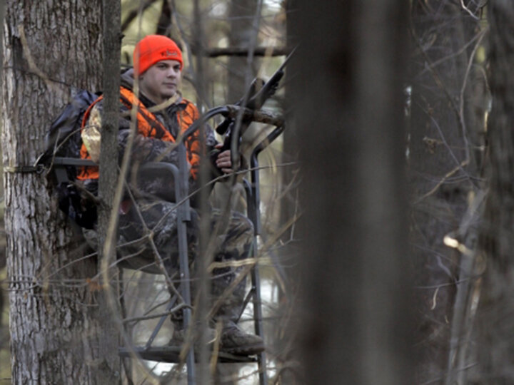 A male hunter sits in a tree stand while wearing camouflage and a hunter orange hat